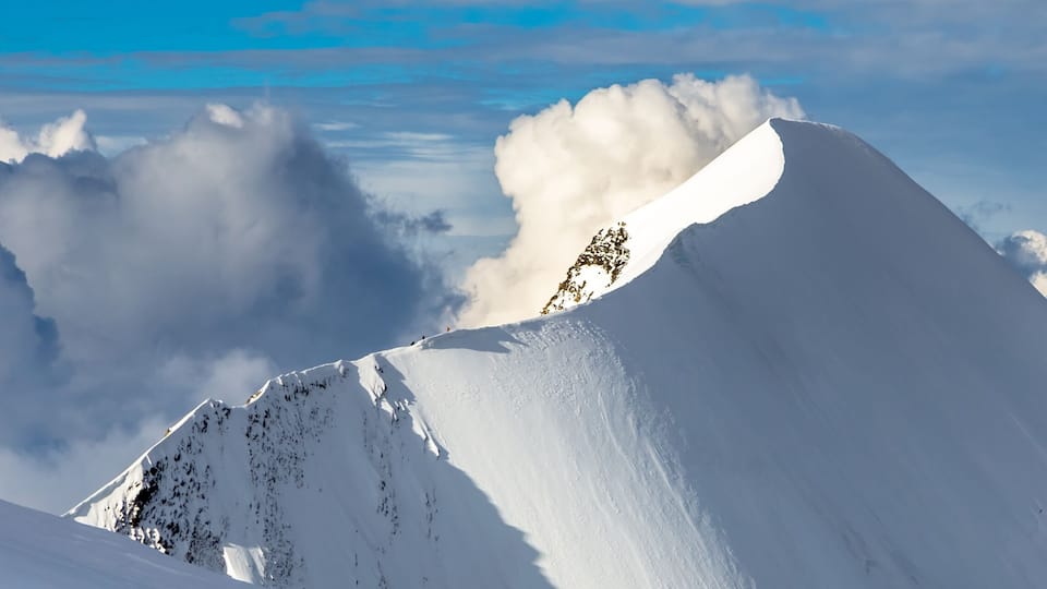 A snow-covered mountain peak with a clear sky and clouds in the background.