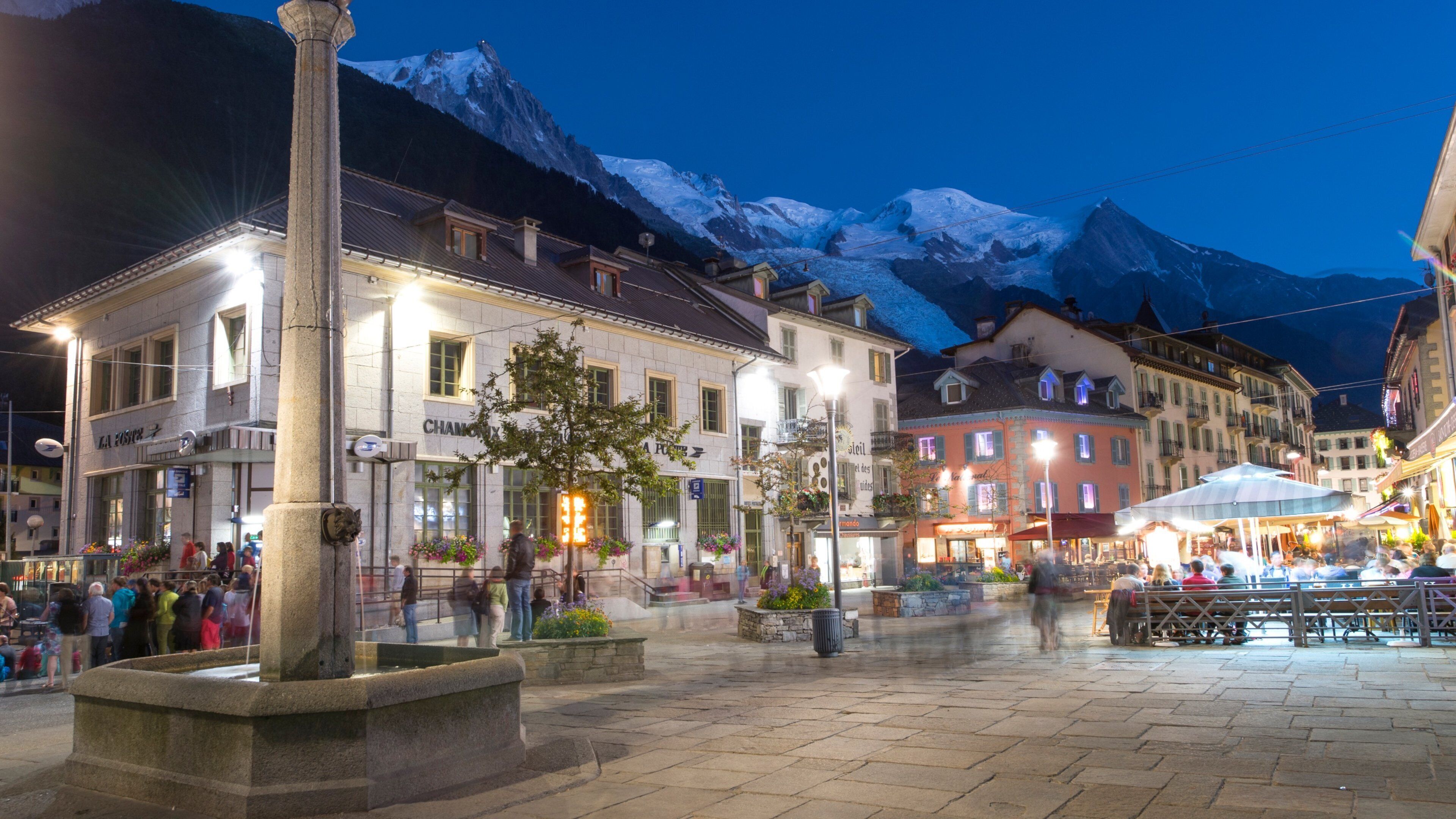 A town square with buildings, a fountain, and snow-capped mountains in the background.
