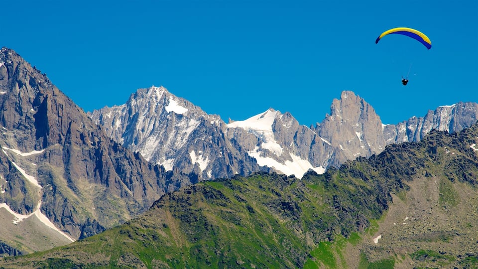 A paraglider flying over a mountainous landscape with snow-capped peaks and lush green valleys.