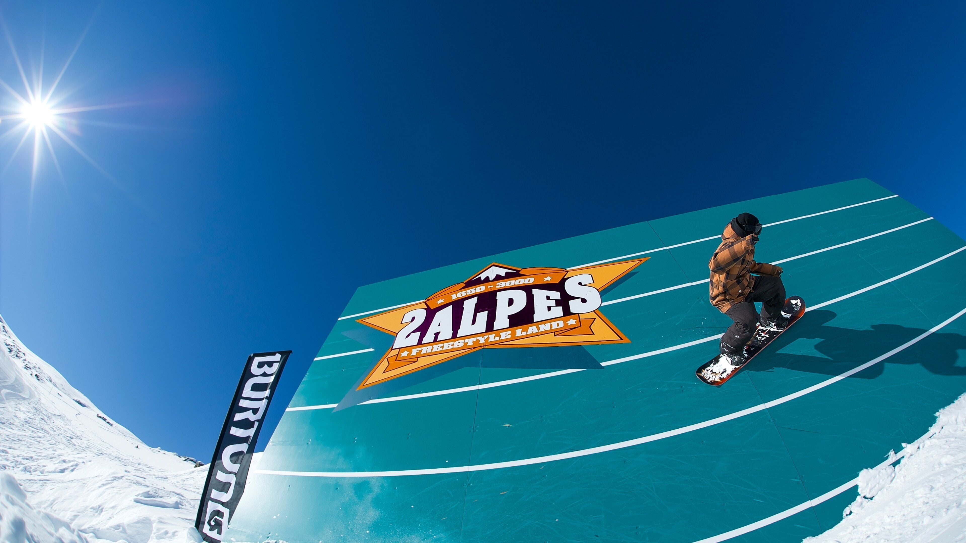 A snowboarder on a blue ramp with a '2ALPES FREESTYLE LAND' logo.