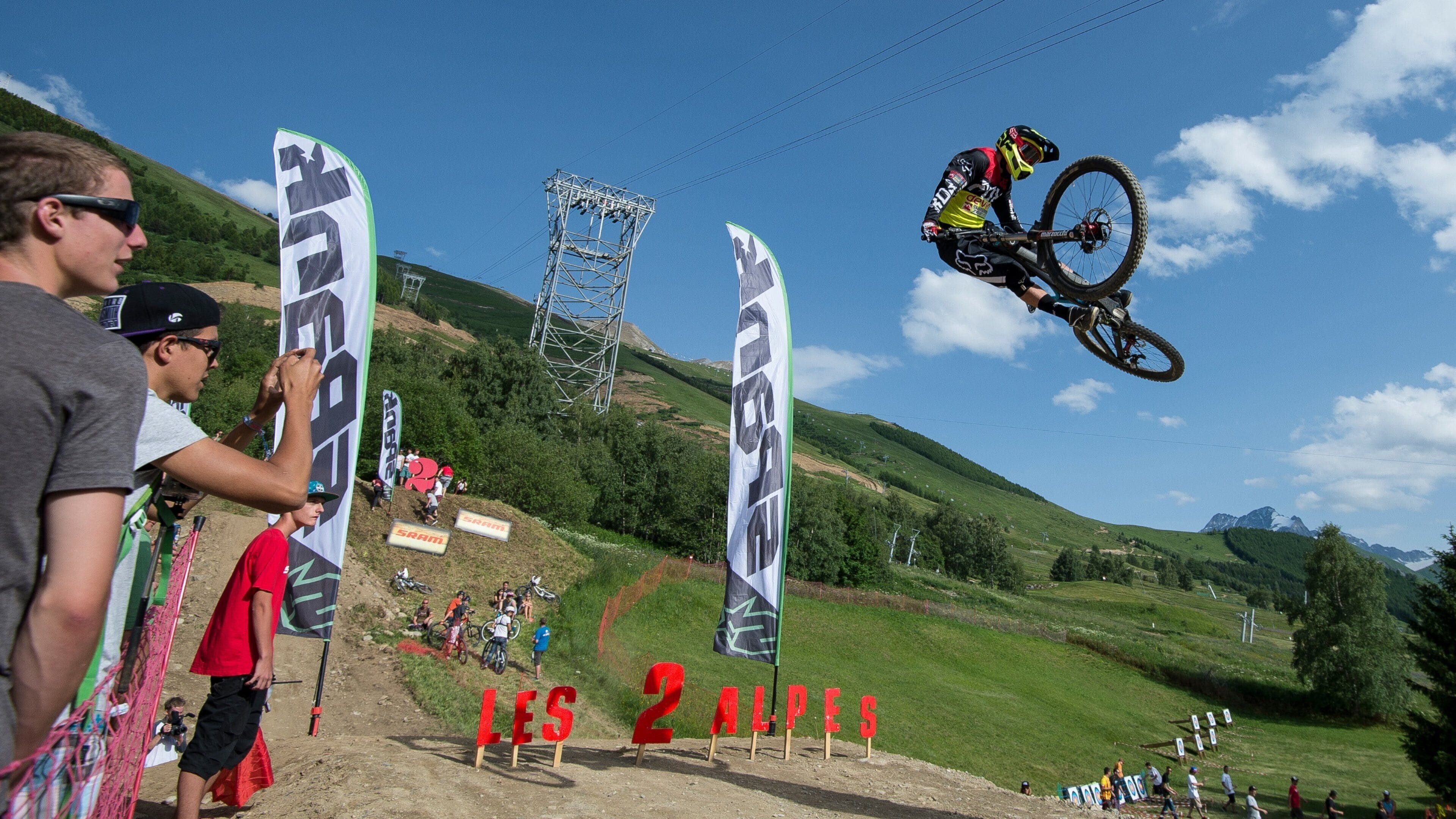 A mountain biking event with a rider performing a jump. Spectators are watching from the sidelines.