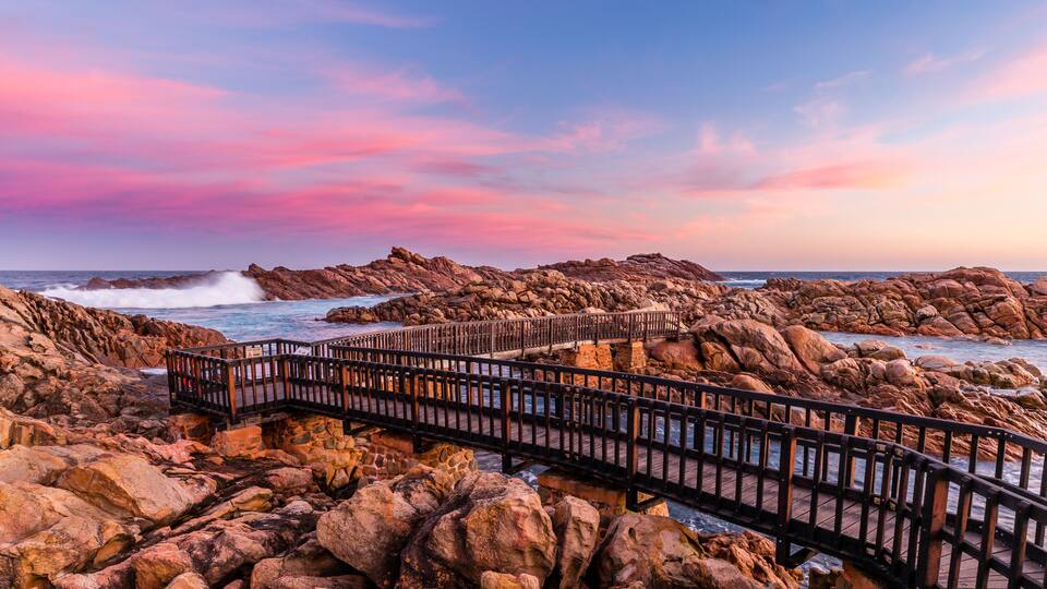 A wooden boardwalk over rocky terrain leading to a coastal area with waves crashing against the rocks.