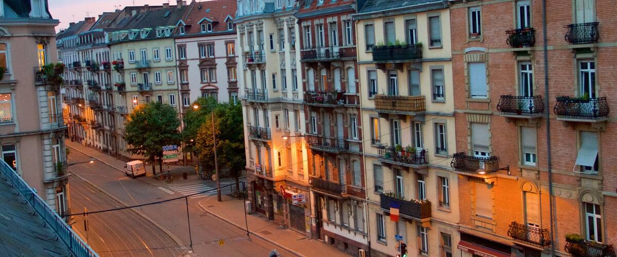 A street lined with multi-story buildings with balconies and awnings, under a twilight sky.