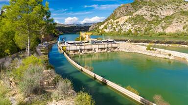 Scenic view of the Xerta Weir (Azud de Xerta) on the Ebro river in Catalonia, Spain, featuring a dam, green water, and rocky hills under a blue sky.