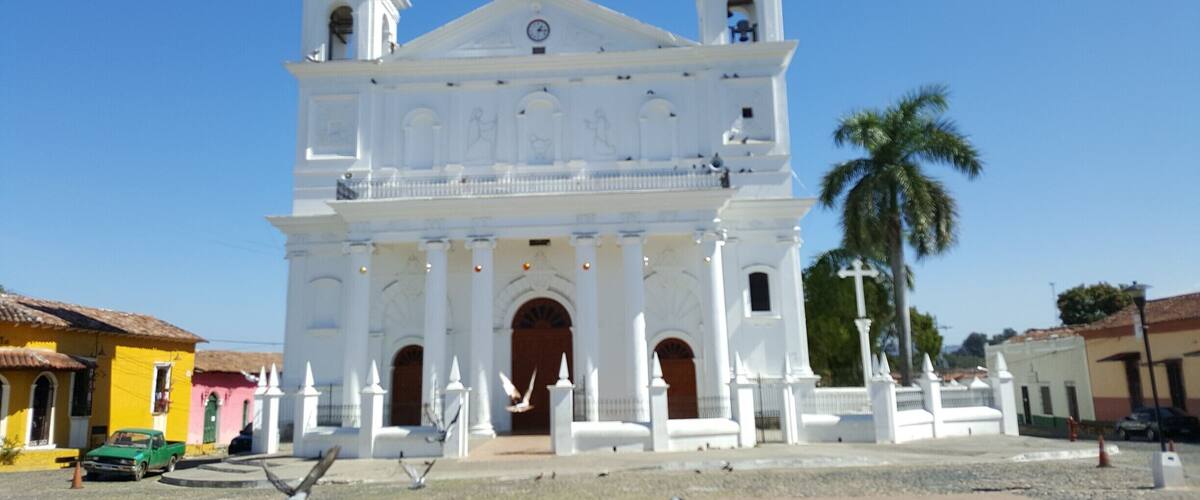 A white church with two bell towers and a central dome, surrounded by palm trees and colorful buildings.