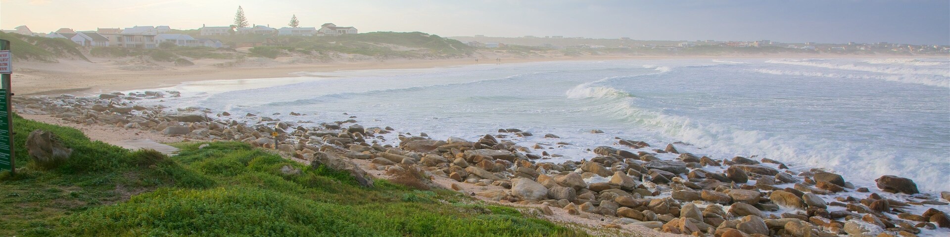 Una playa rocosa con olas rompiendo en la orilla, un área con césped en primer plano y un pueblo distante en el horizonte.