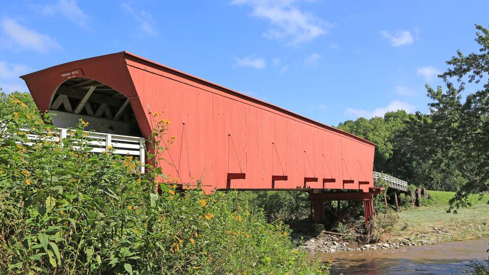 A red covered bridge over a river with greenery and a clear blue sky.