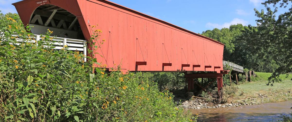 A red covered bridge over a river with greenery and a clear blue sky.