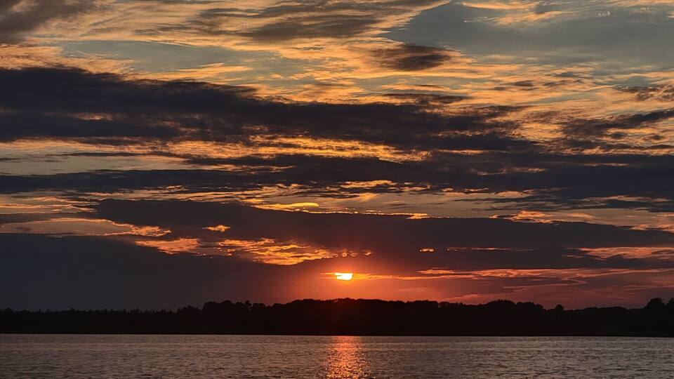 A sunset over a body of water with a silhouette of trees and a cloudy sky.
