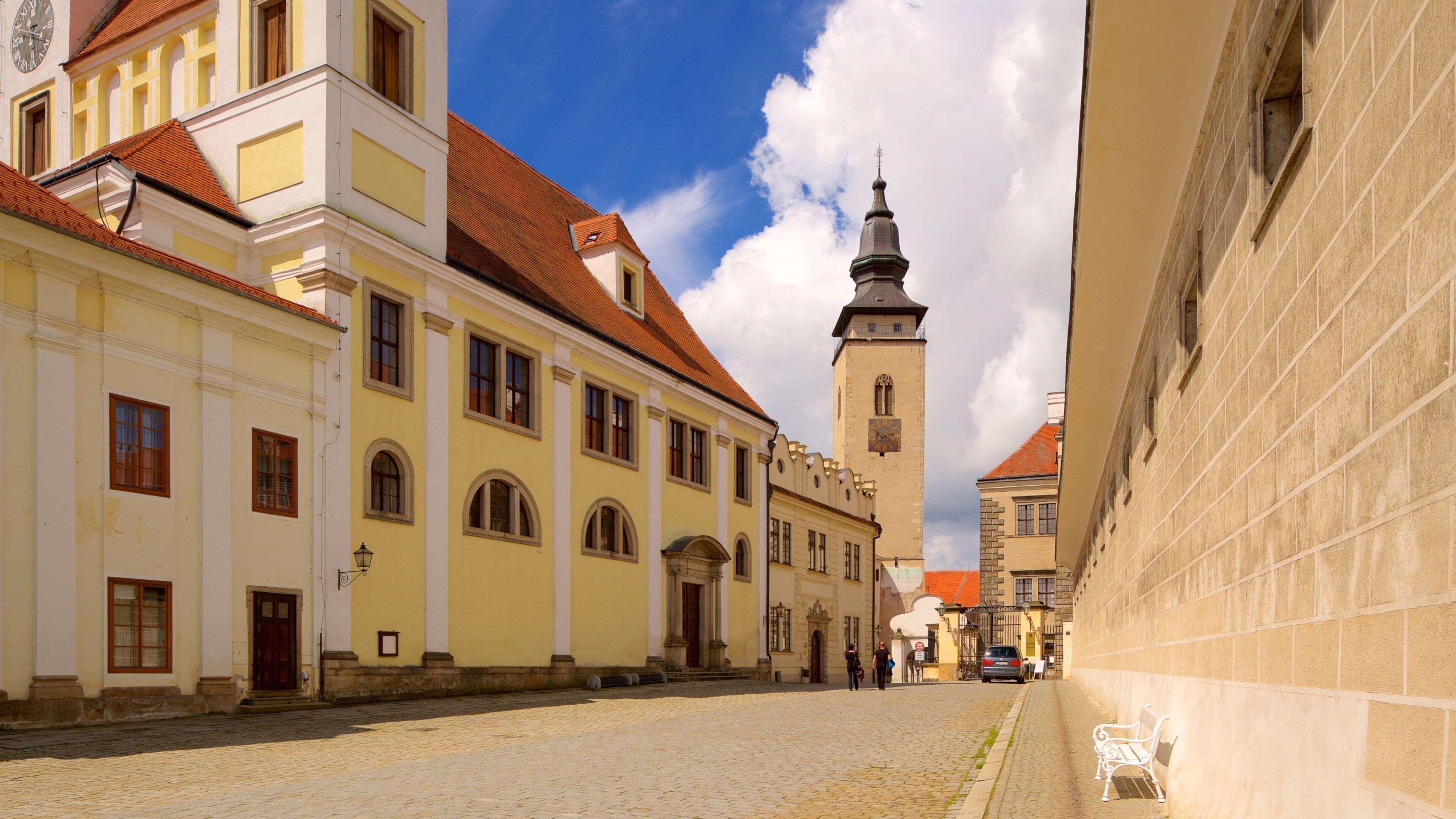 A cobblestone street lined with historic buildings, a prominent church tower, and a bench.