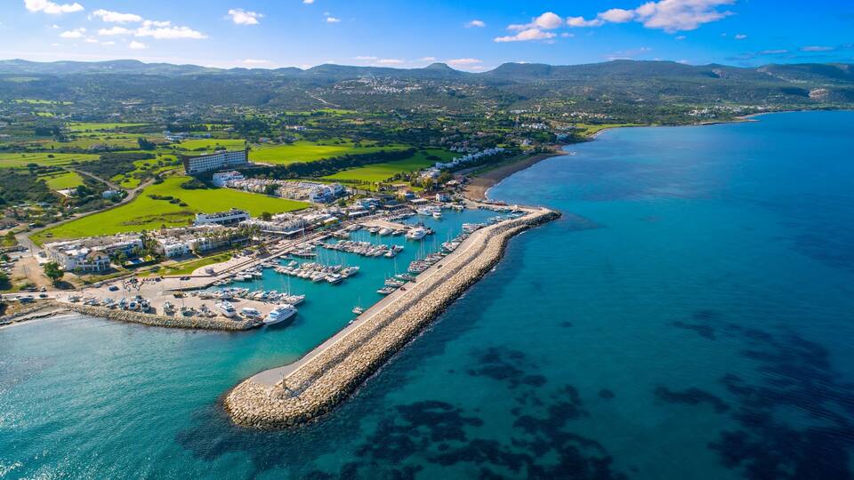 An aerial view of a coastal town with a marina, a long pier, and surrounding green hills.