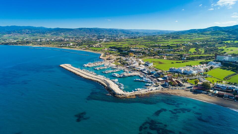An aerial view of a coastal town with a marina, green hills, and a clear blue sea.