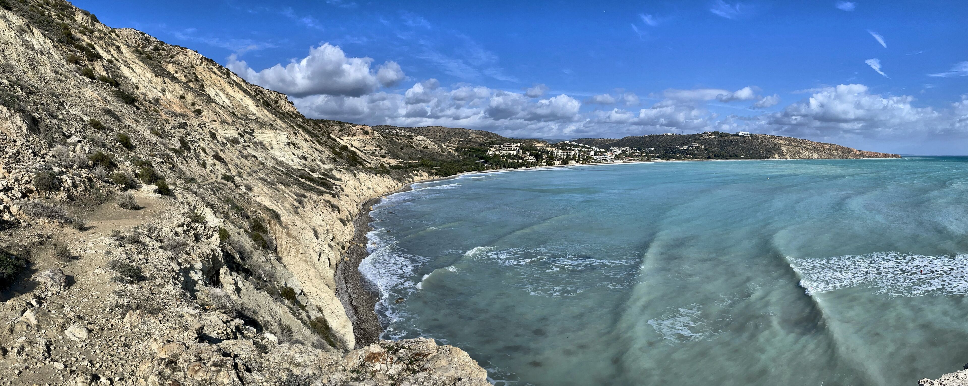 A coastal landscape with a rocky cliff, clear turquoise waters, and a distant hill with buildings.
