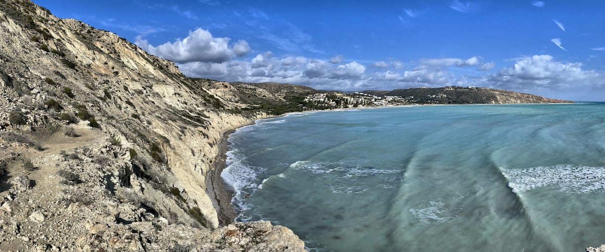 A coastal landscape with a rocky cliff, clear turquoise waters, and a distant hill with buildings.
