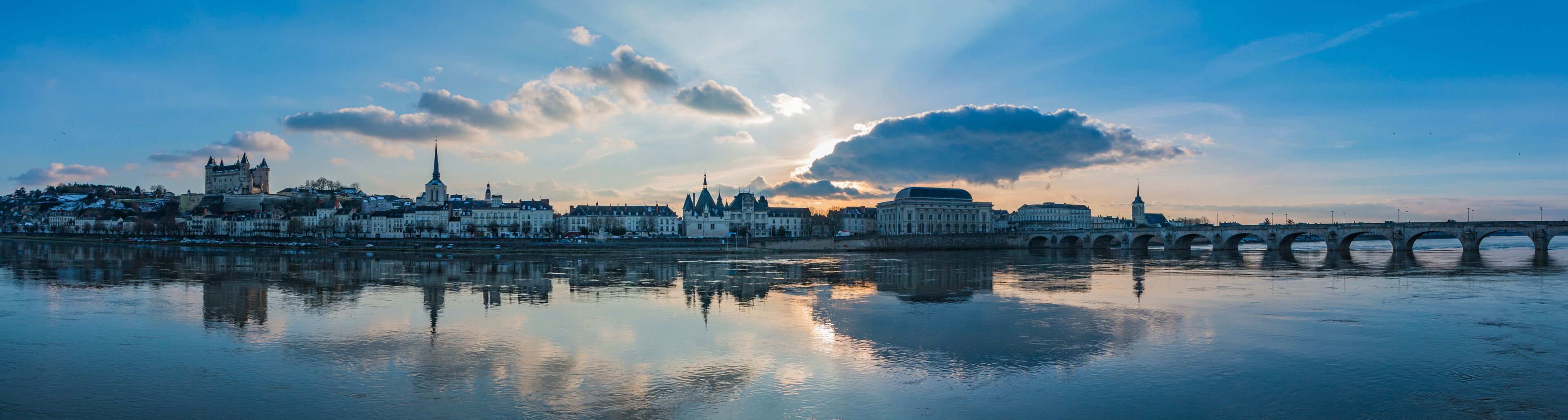 Une vue sur les toits d’une ville, avec un château imposant, une cathédrale et un pont enjambant une rivière paisible.