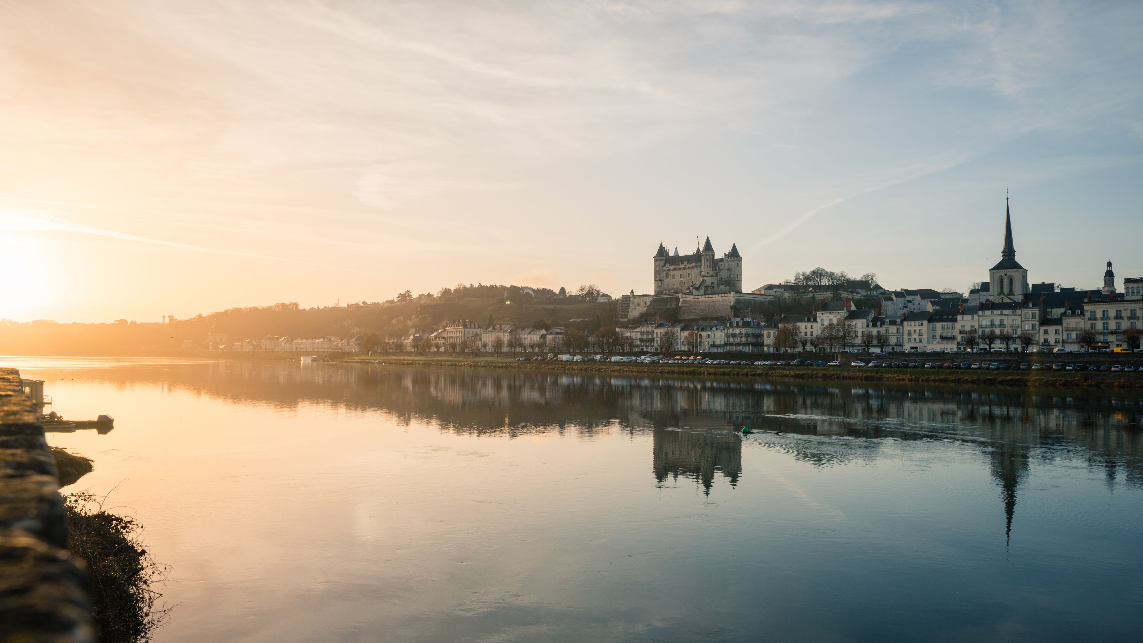 Um rio calmo refletindo uma cidade histórica com um castelo e igreja imponentes.