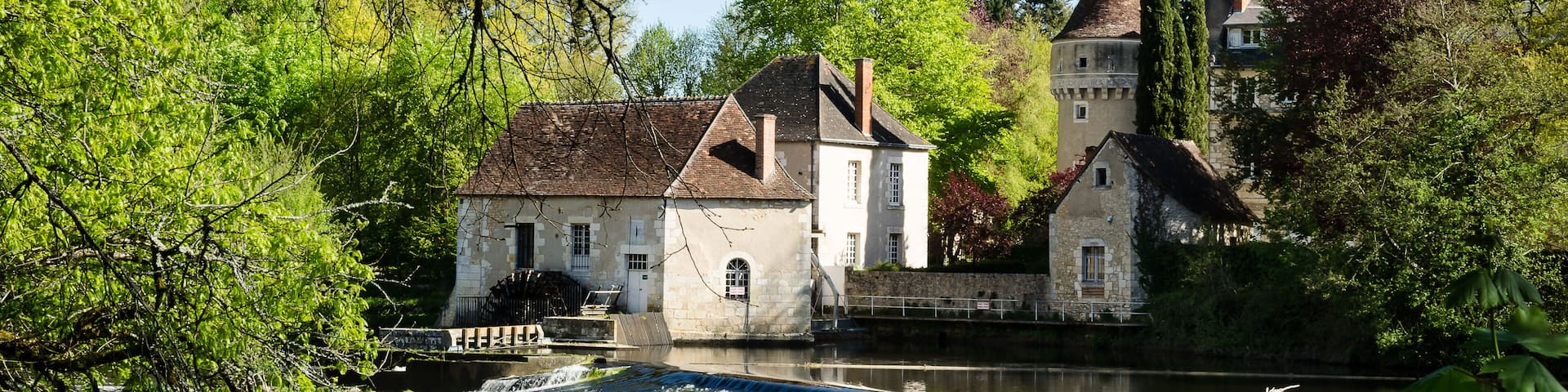 A historic building with a water mill, surrounded by lush greenery and a calm river.