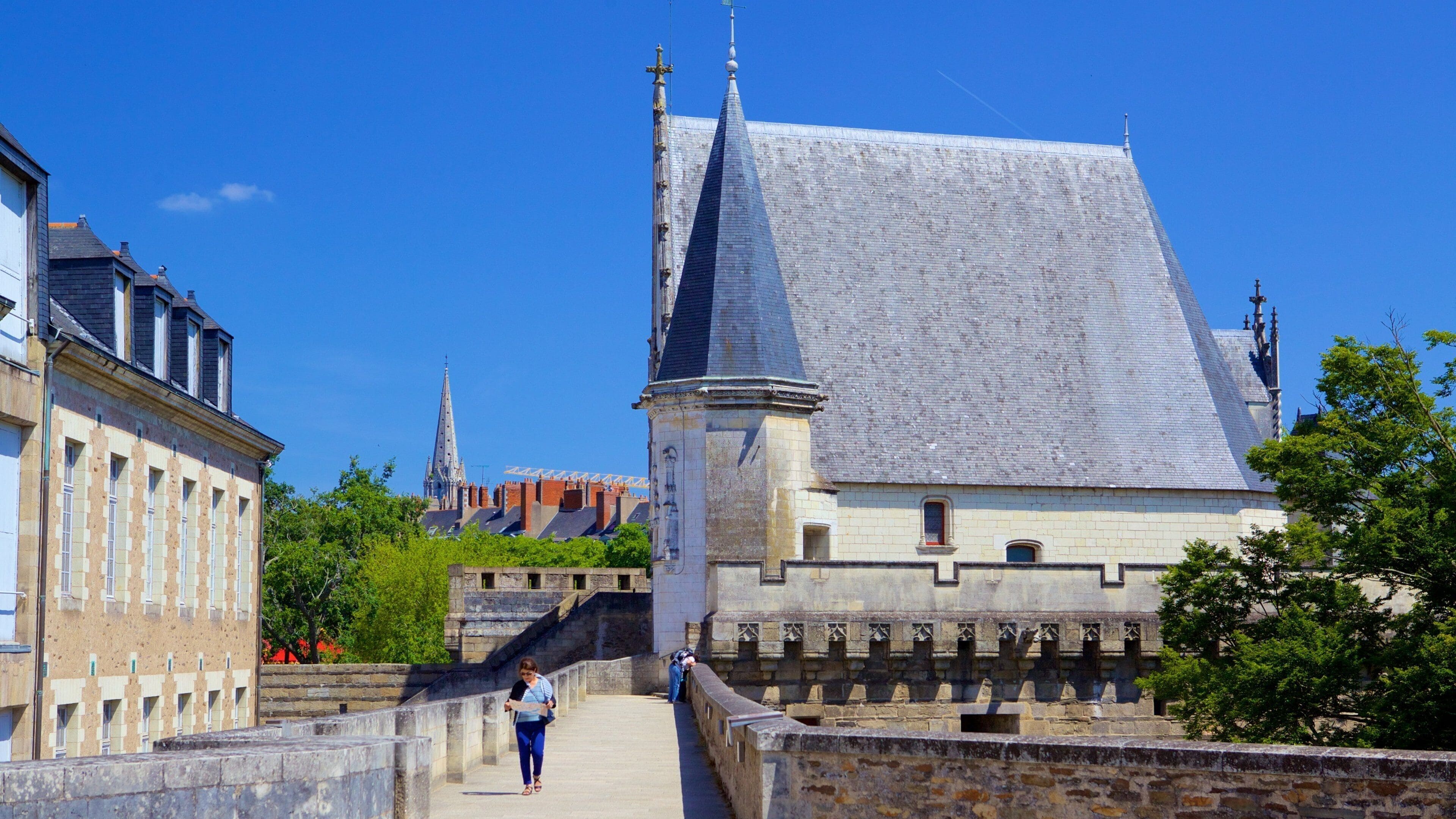 A historic building with a steeply pitched roof and a tall spire, surrounded by stone walls and greenery.
