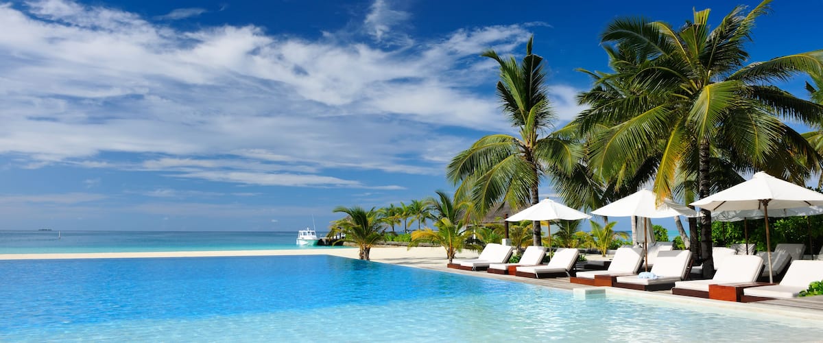 A clear blue pool with white lounge chairs and palm trees under a bright blue sky.