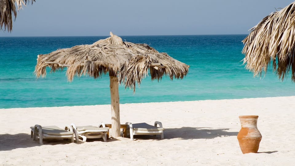 A beach with a thatched umbrella, lounge chairs, and a clay pot.