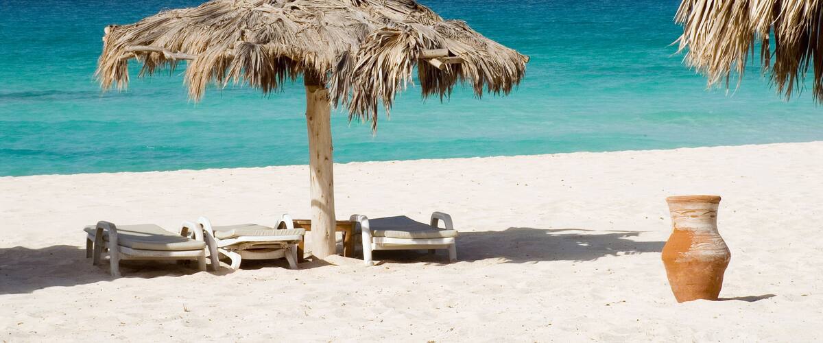 A beach with a thatched umbrella, lounge chairs, and a clay pot.