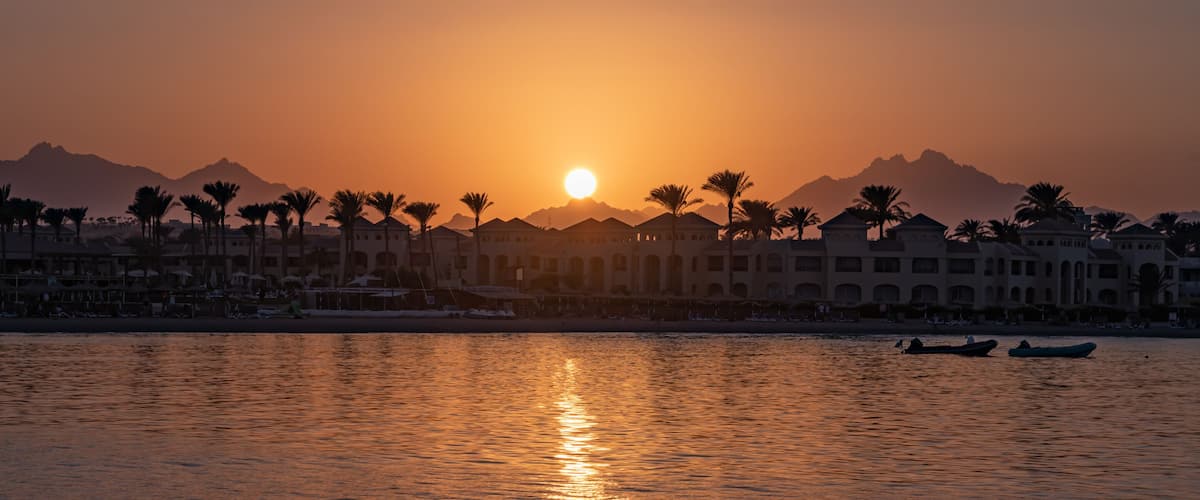 A sunset over a coastal city with palm trees, a beach, and mountains in the background.
