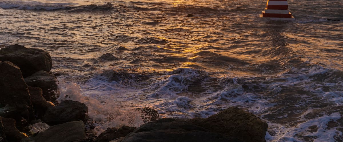 A lighthouse at sunset with waves crashing against rocks.