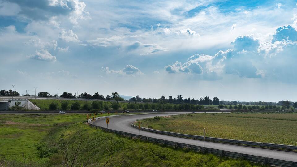 A curved highway with guardrails, surrounded by green fields and a cloudy sky.