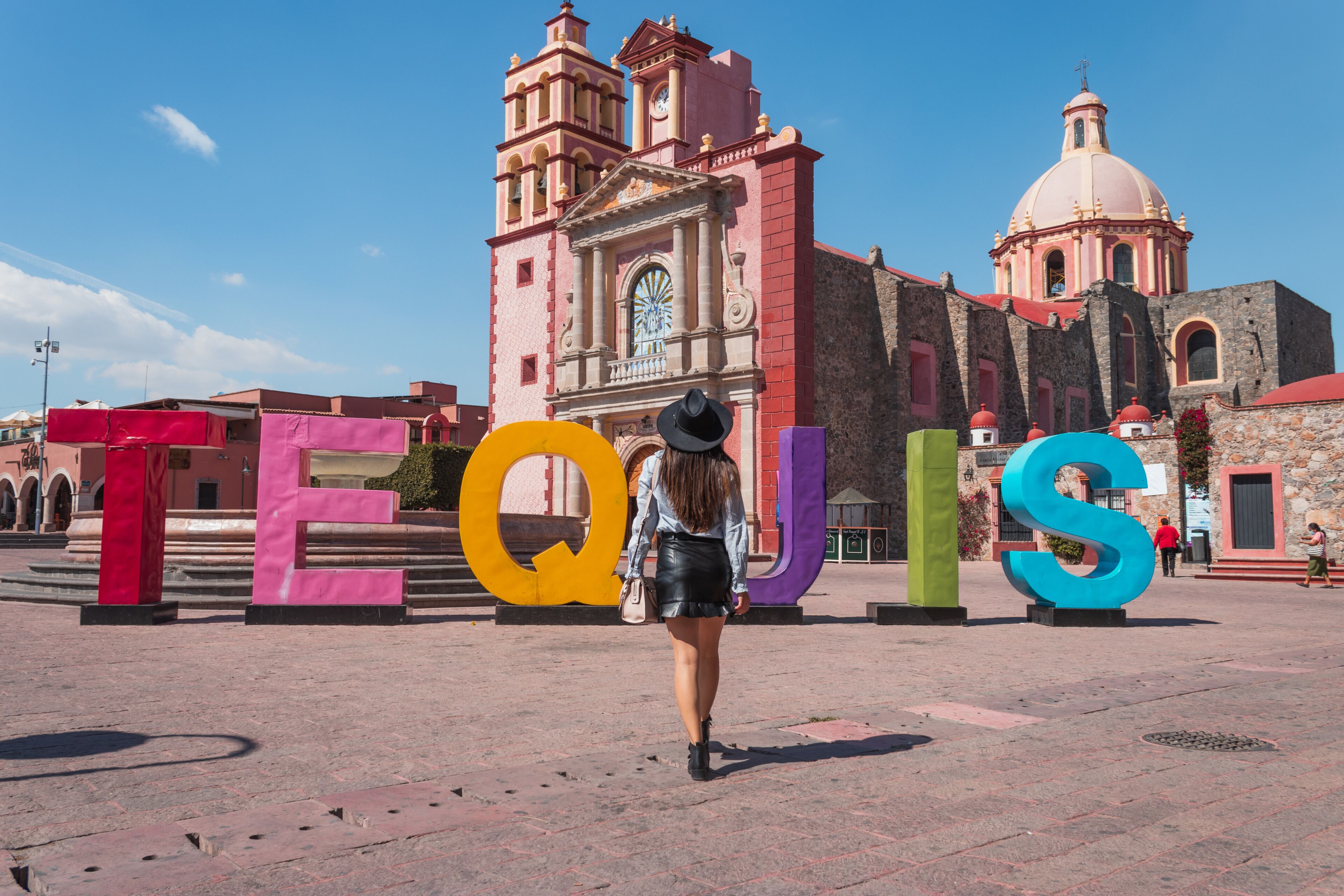 A woman walking past large colorful letters spelling 'TEQUIS' in front of a historic church with a bell tower and dome.