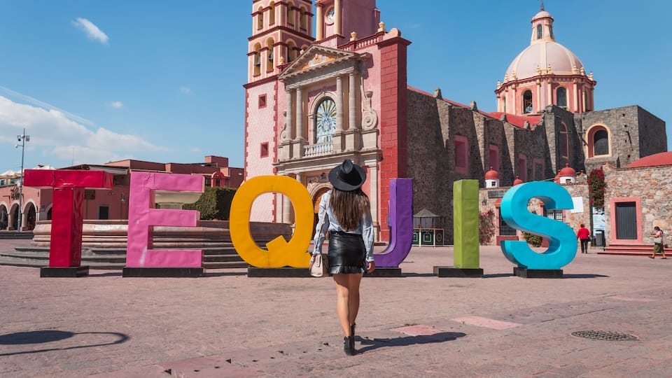 A woman walking past large colorful letters spelling 'TEQUIS' in front of a historic church with a bell tower and dome.