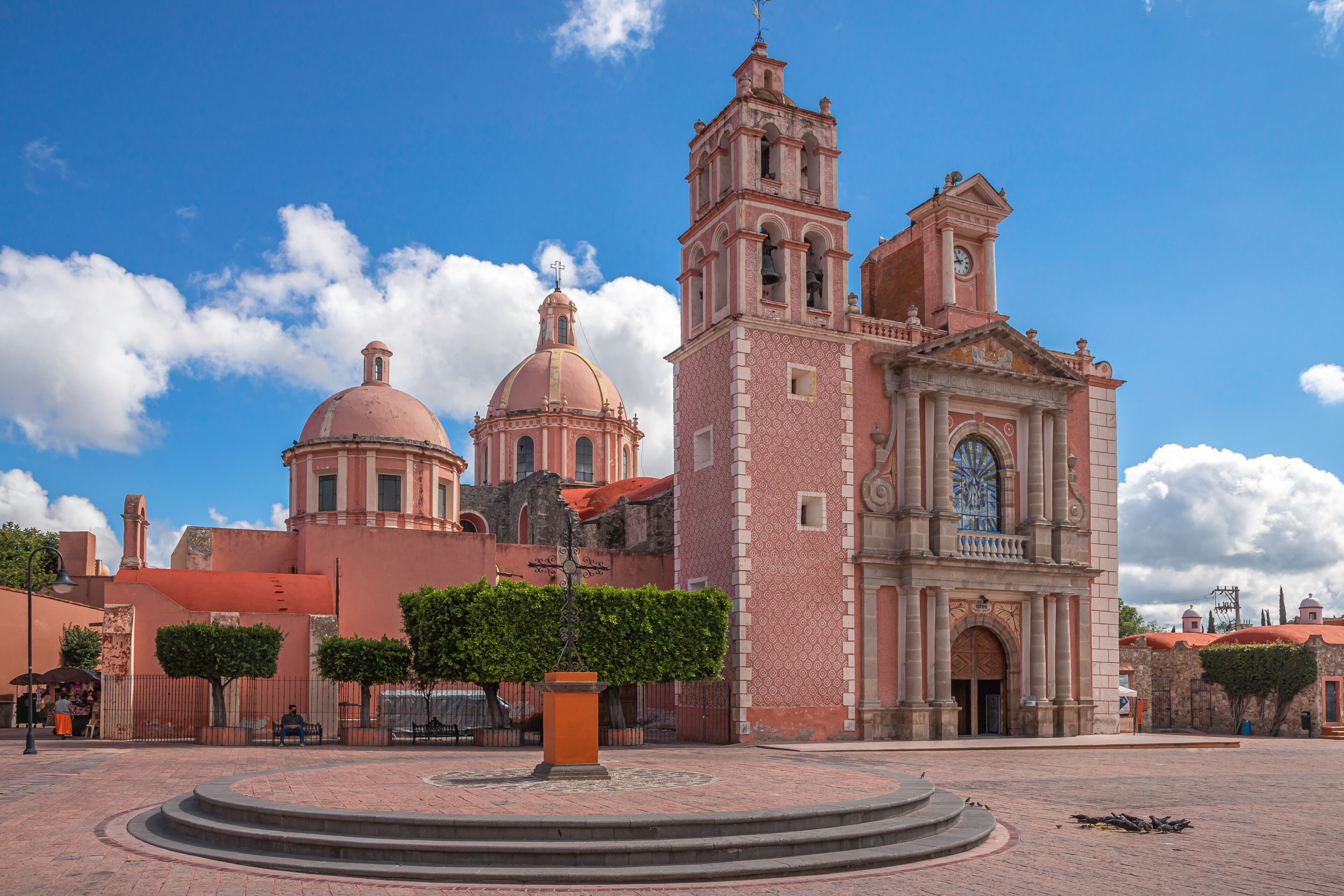 A historic church with two domes and a bell tower, surrounded by a plaza with a central fountain and trimmed hedges.