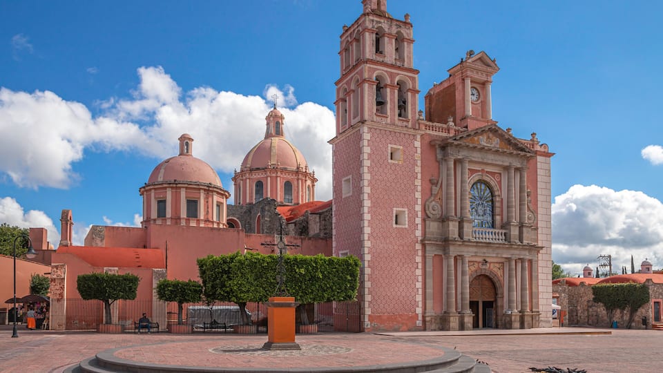 A historic church with two domes and a bell tower, surrounded by a plaza with a central fountain and trimmed hedges.