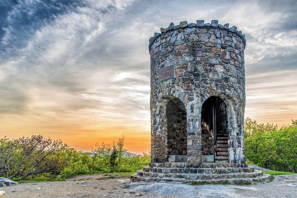 A stone tower with arched openings and a staircase leading up to a door, surrounded by greenery and a cloudy sky.