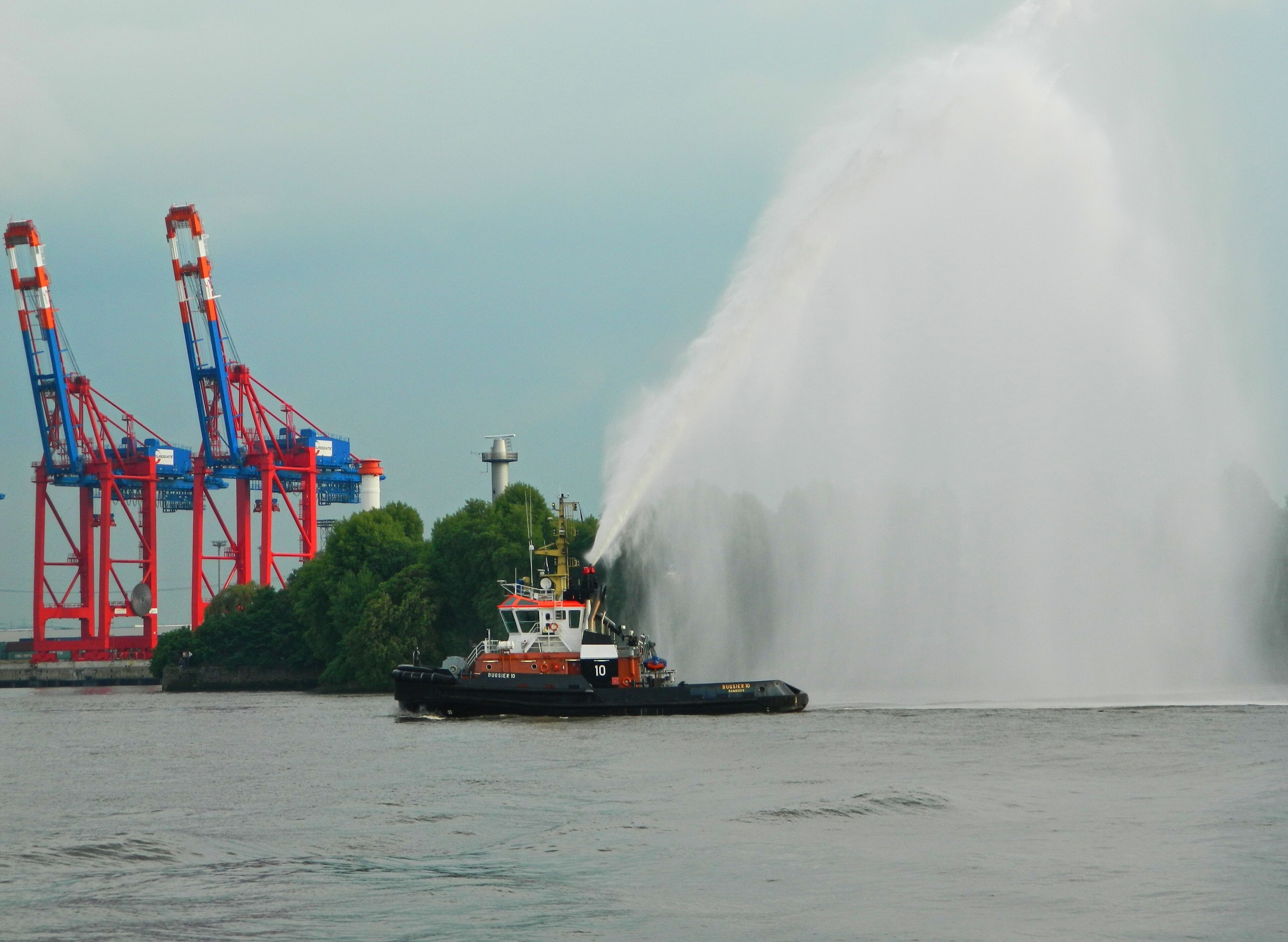 A tugboat named 'Burgasier D' with a water jet in the foreground, a crane, and a lighthouse in the background.