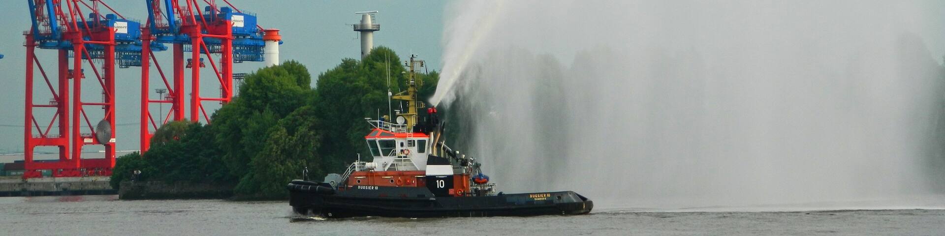 A tugboat named 'Burgasier D' with a water jet in the foreground, a crane, and a lighthouse in the background.