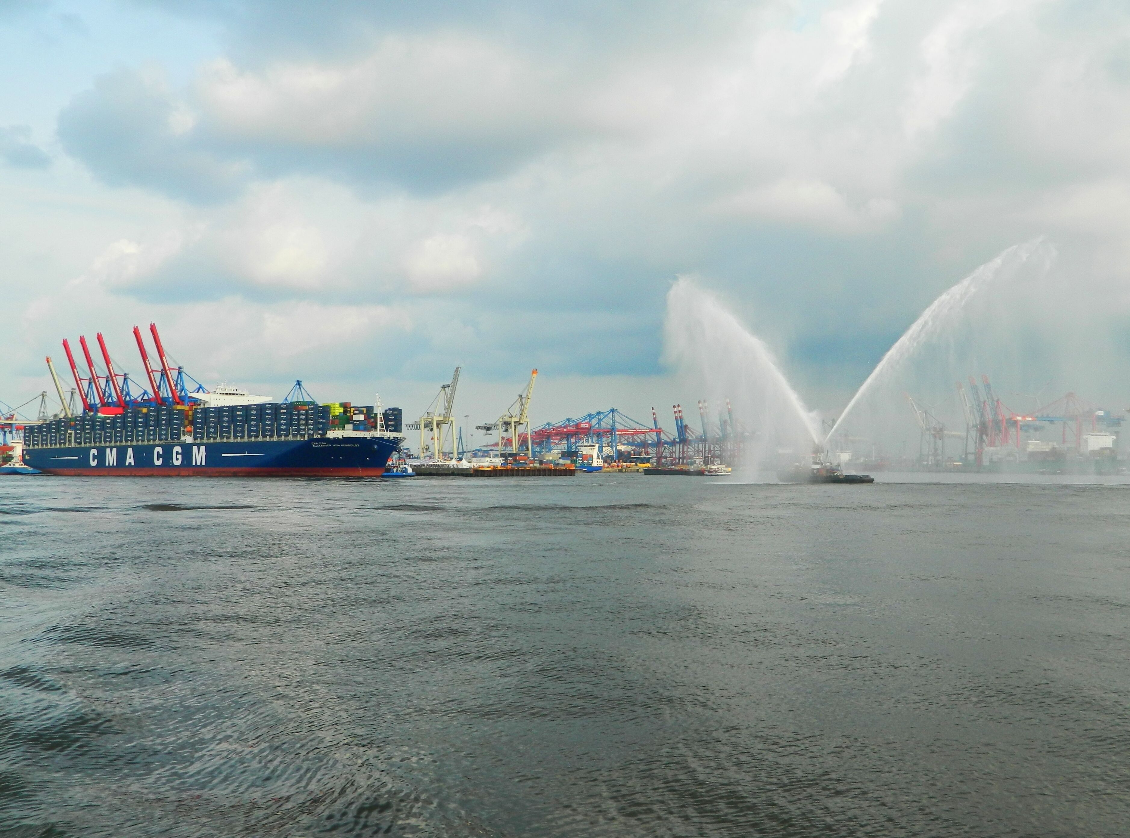 A cargo ship with the letters "CMA CGM" on its side, docked at a port with cranes and a water fountain in the background.