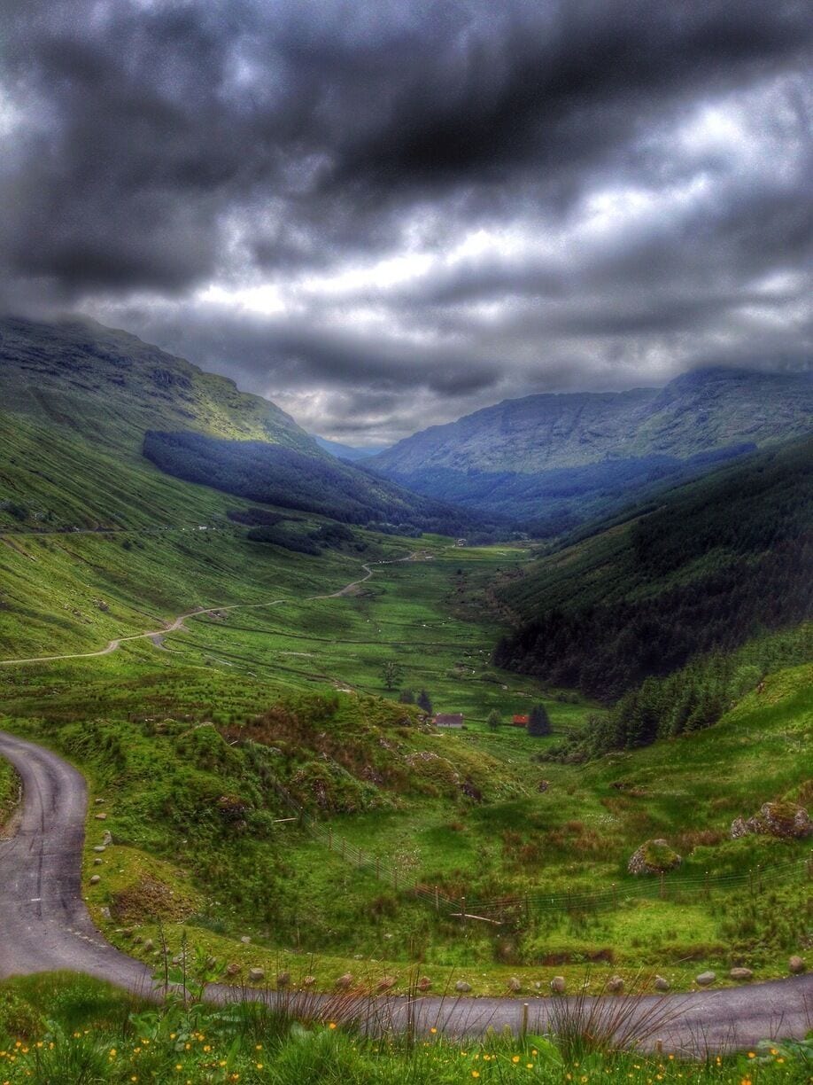 A lush green valley with a winding road, surrounded by hills and under a cloudy sky.