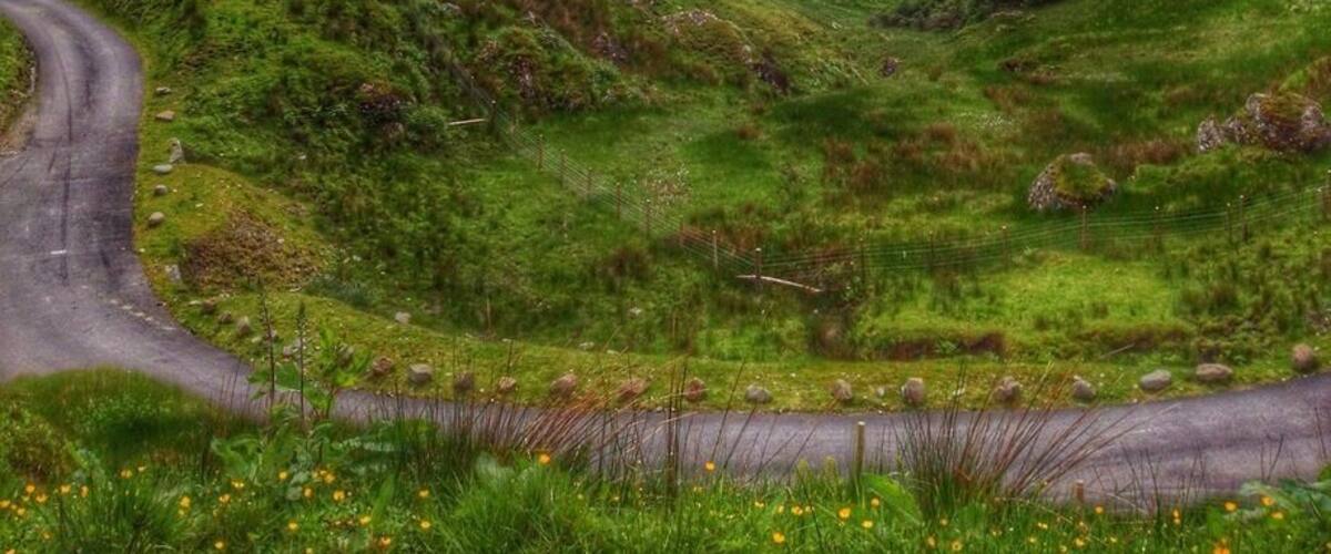 A lush green valley with a winding road, surrounded by hills and under a cloudy sky.
