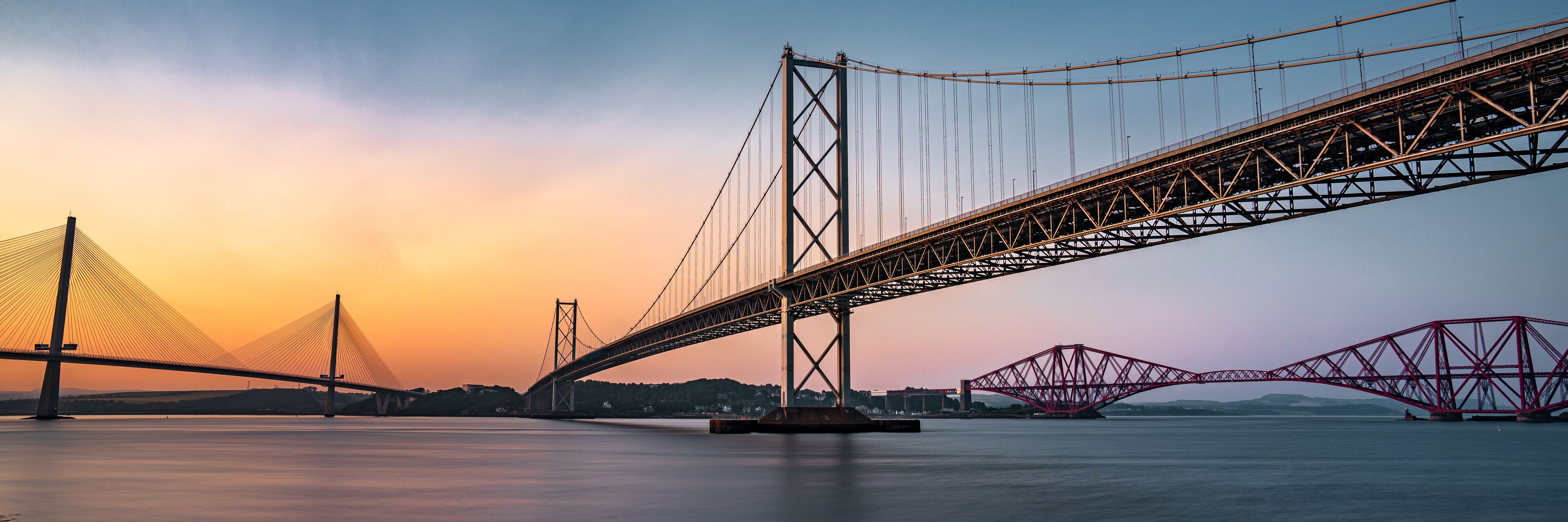 A suspension bridge with cables and towers, a red truss bridge, and a calm body of water.