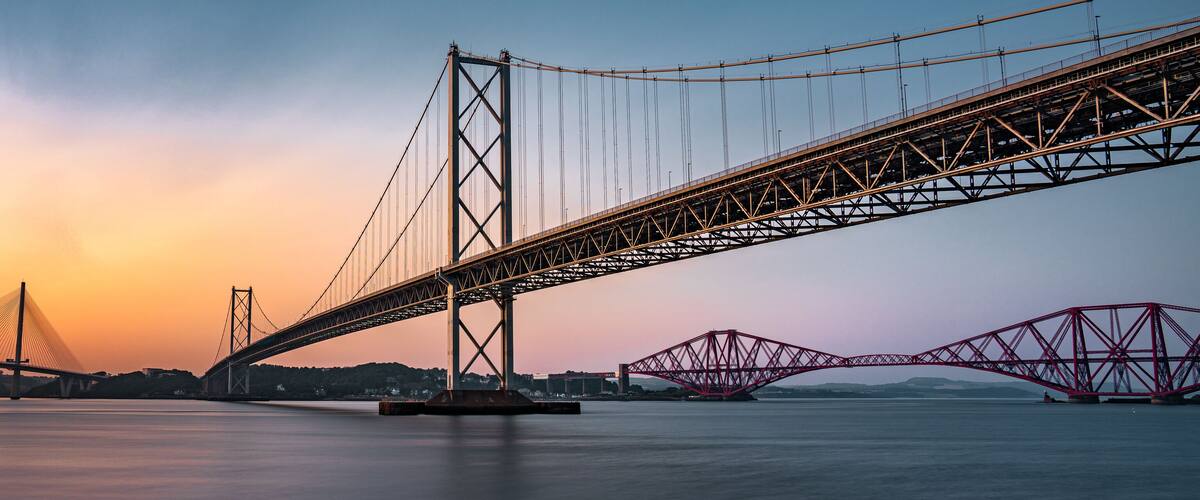 A suspension bridge with cables and towers, a red truss bridge, and a calm body of water.