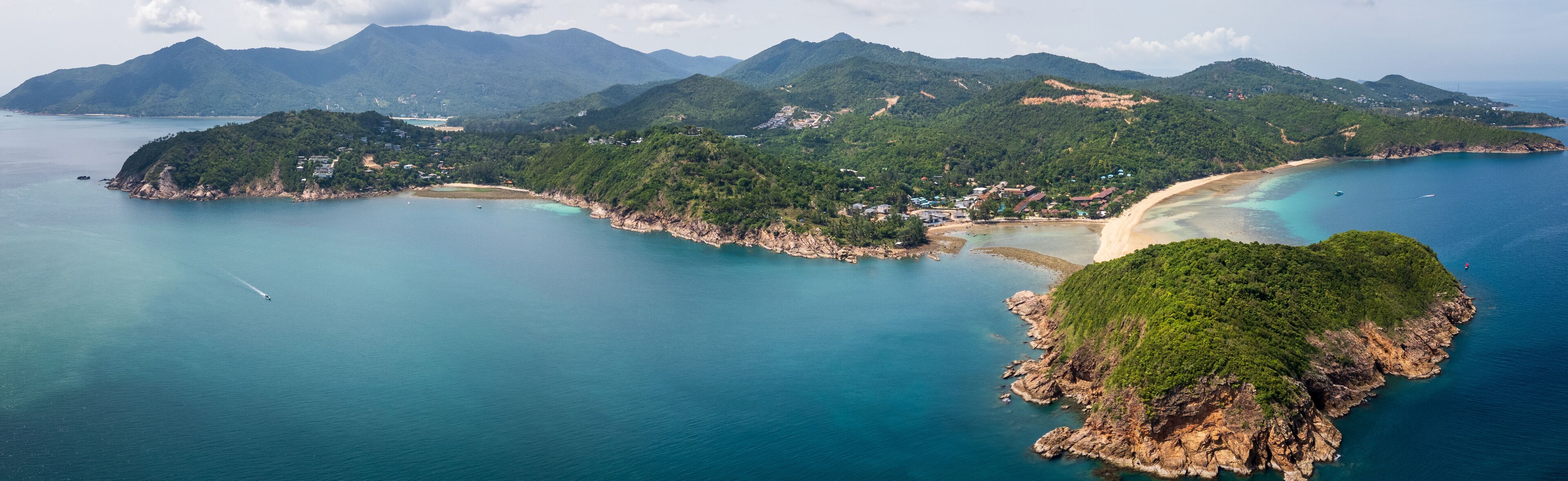 Vista aérea de una zona costera con playa de arena, aguas turquesas cristalinas y colinas verdes exuberantes.