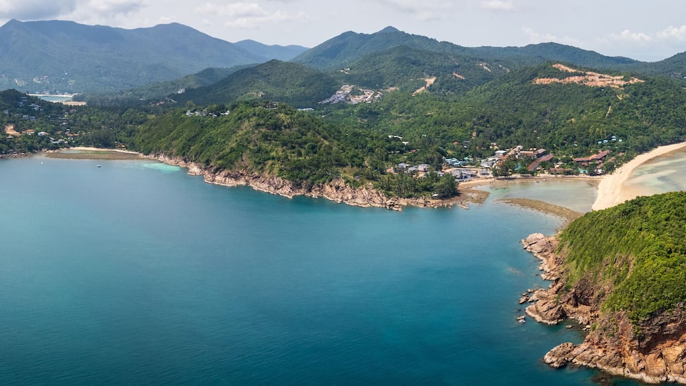 An aerial view of a coastal area with a sandy beach, clear turquoise waters, and lush green hills.