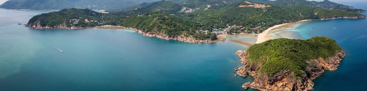An aerial view of a coastal area with a sandy beach, clear turquoise waters, and lush green hills.