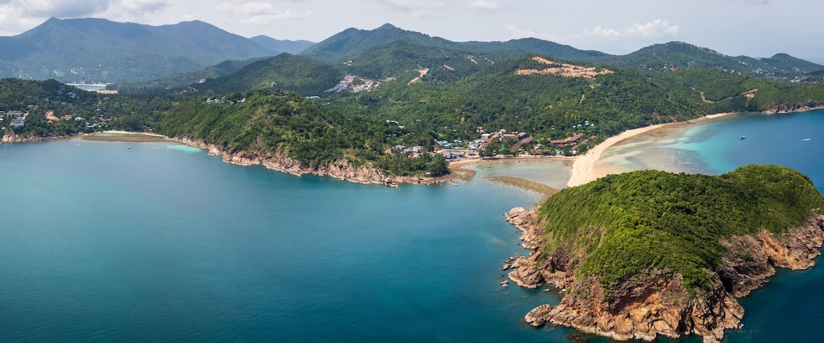 Une vue aérienne d’une zone côtière avec une plage de sable, des eaux turquoise et limpides, et des collines verdoyantes.