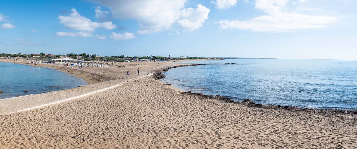 A sandy beach with a stone structure, calm waters, and a distant shoreline with buildings.