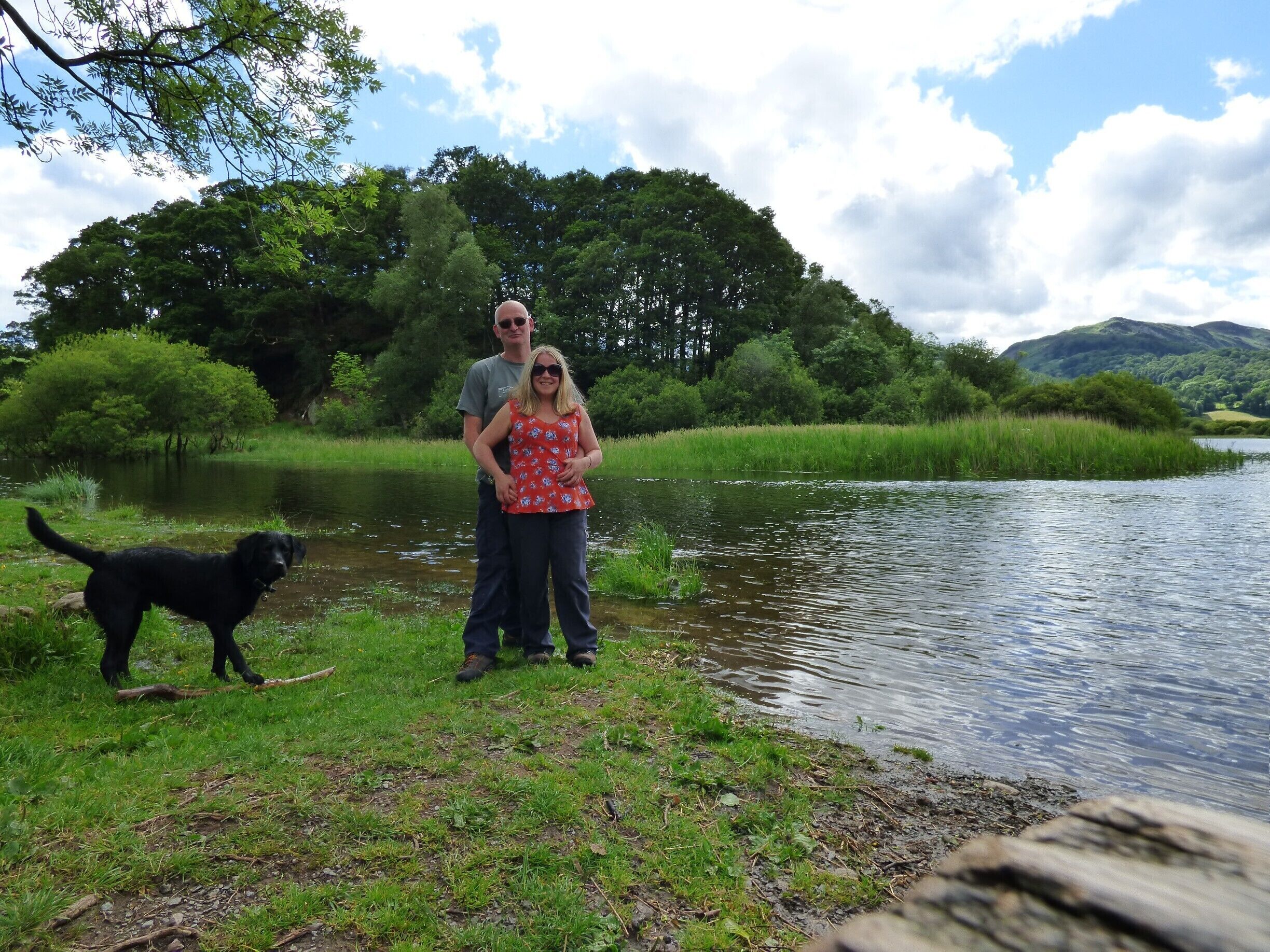 A couple standing by a calm river with a dog, surrounded by lush greenery and trees.