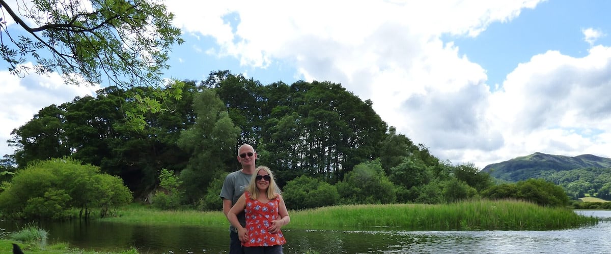 A couple standing by a calm river with a dog, surrounded by lush greenery and trees.