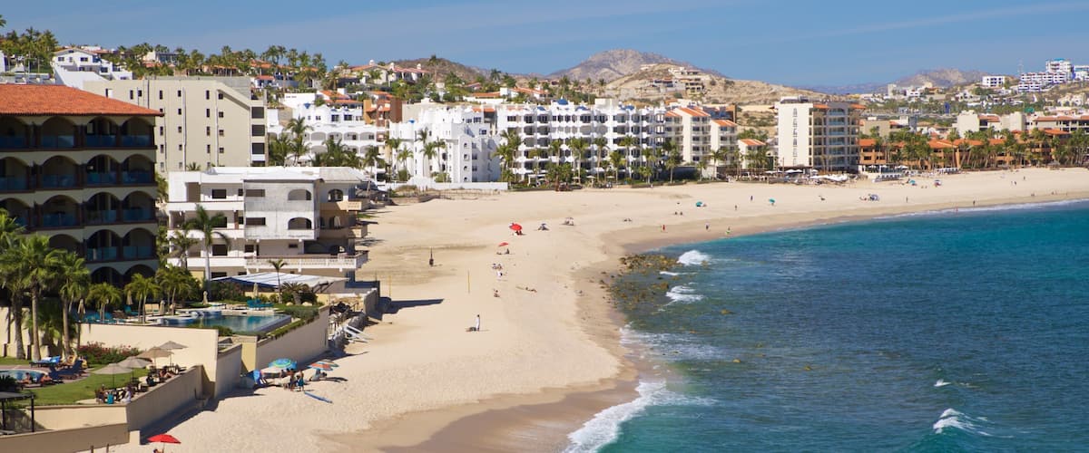A coastal resort with a sandy beach, clear blue waters, and a backdrop of mountains and residential buildings.