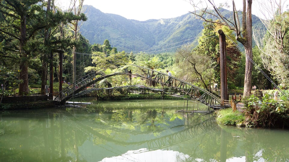 A wooden bridge over a calm river, surrounded by lush greenery and mountains in the background.
