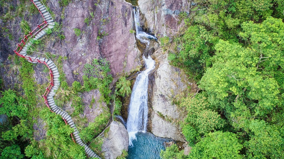 A waterfall cascading down a rocky cliff, surrounded by lush greenery and a winding red path.